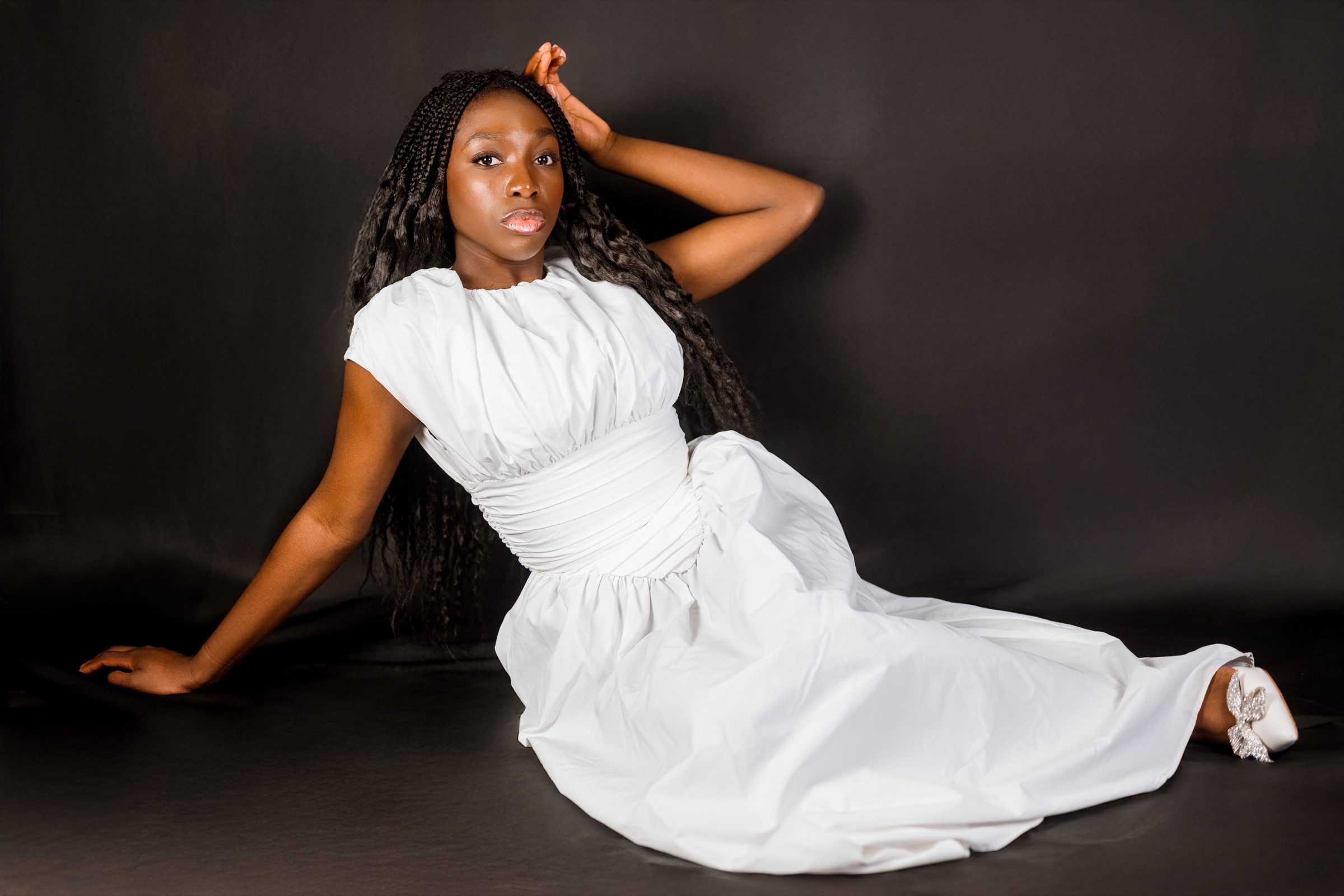 White gown on a black studio floor, one hand in her braids