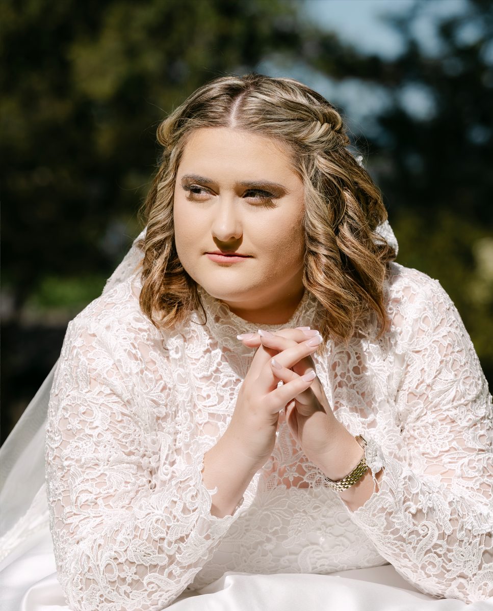 Bride in lace, hands clasped, sunlit lawn behind