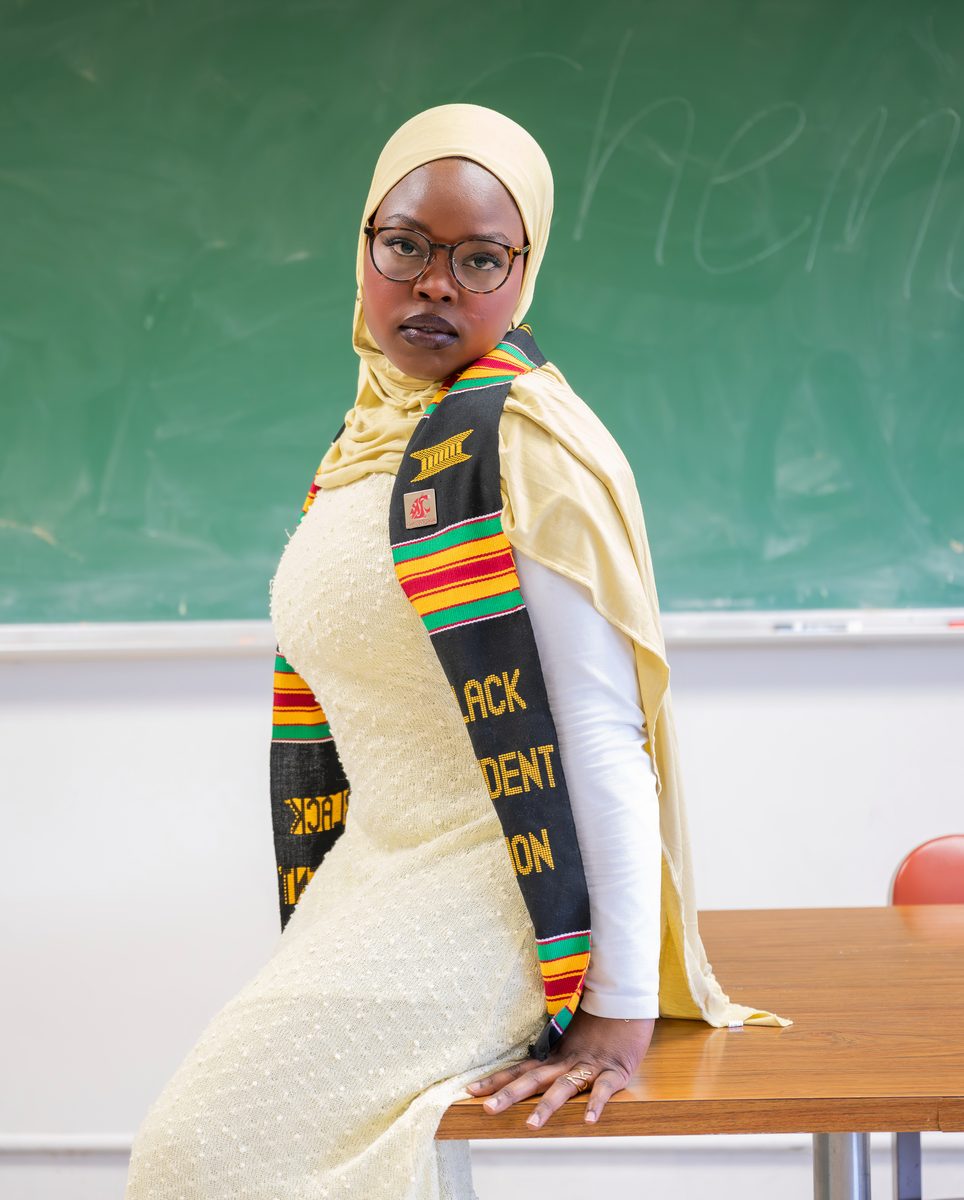 Graduate in a kente stole, seated on a desk before a chalkboard