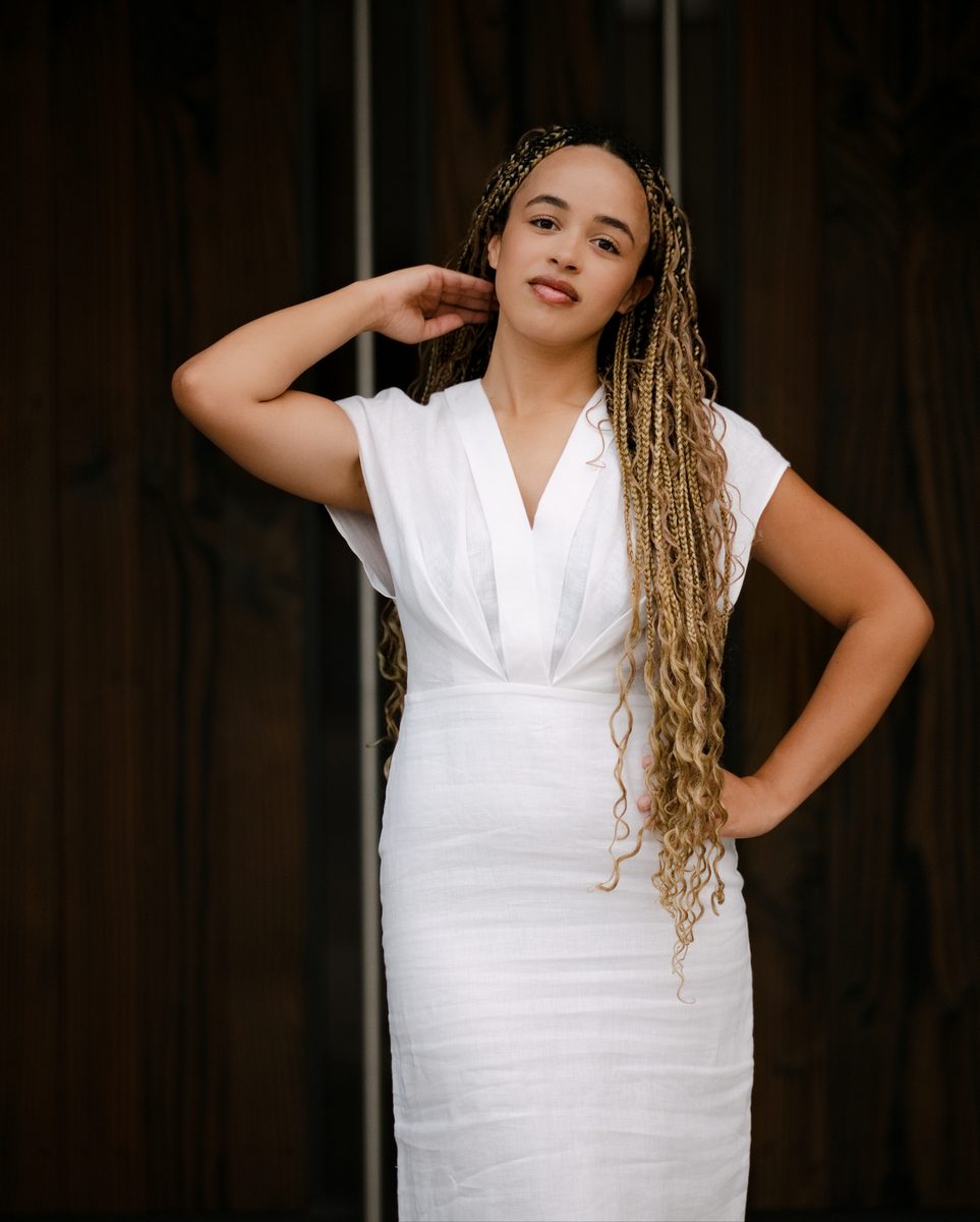 White linen dress against dark wood, hand in her braids