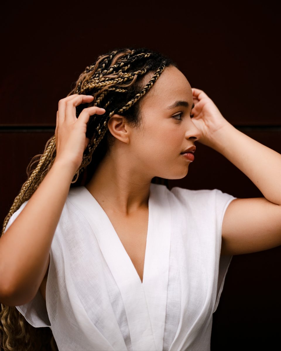 White linen dress, hands in braided hair, dark wood behind