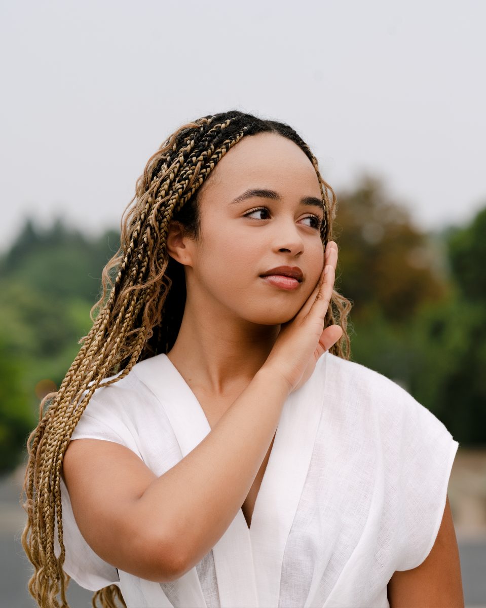 White linen, hand at her cheek, green trees behind